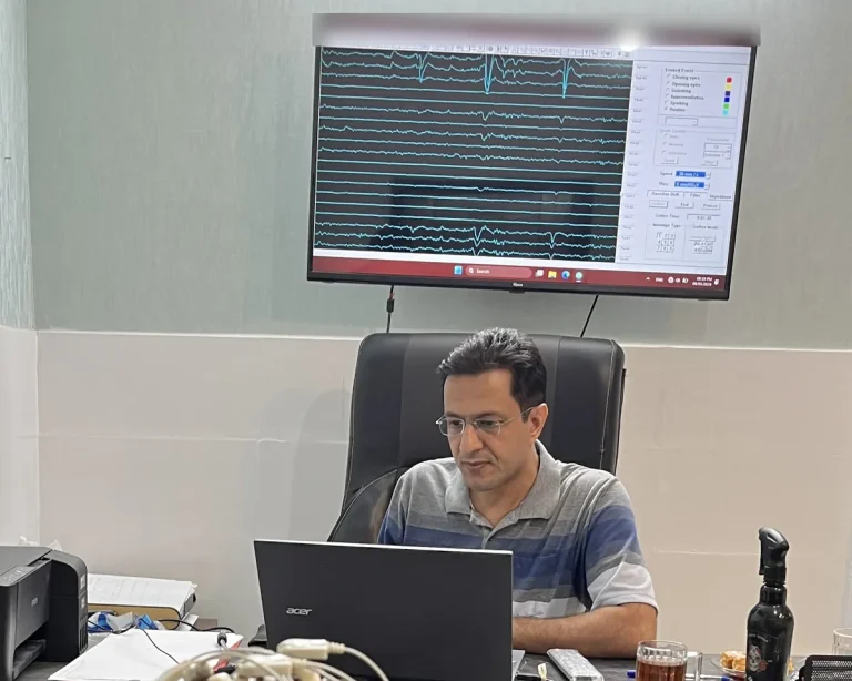 دکتر داود صمدی، دکترای تخصصی علوم اعصاب، پشت میز کار در کلینیک مشهد در حال بررسی امواج مغزی (EEG) روی نمایشگر.